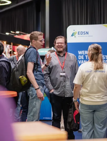 Drie mensen in gesprek op de drukbezochte J-Spring 2025, waarbij een vrouw een t-shirt met het Flink Software logo draagt.