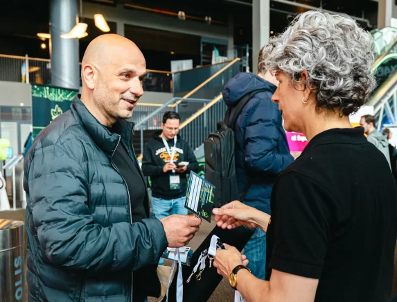 Adis Ćorović in een zwarte jas in gesprek met een vrouw met grijs, krullend haar, terwijl zij hem een badge en een keycord overhandigt op J-Spring 2026.