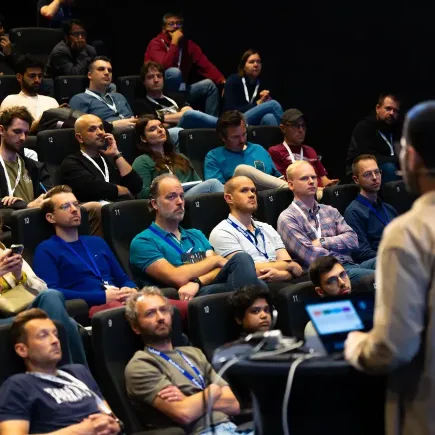 Adis Ćorović zit in het publiek in een donkere zaal met theaterstoelen en luistert aandachtig naar een presentatie van een spreker op J-Spring 2026.