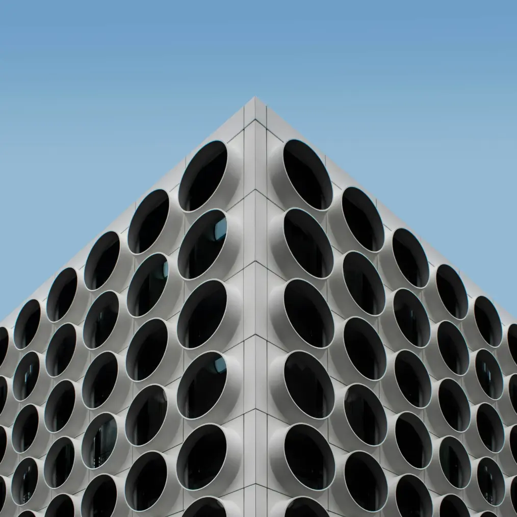 Architecturale close-up van de hoek van een modern, wit gebouw met een patroon van grote, ronde, diepliggende ramen tegen een strakblauwe lucht.