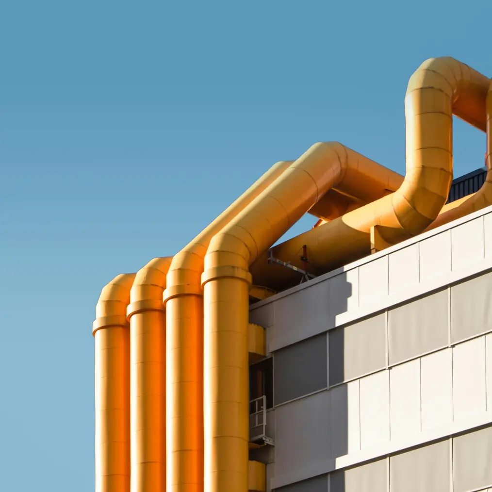 Architectonisch detail van grote, gele pijpleidingen aan de zijkant van een modern gebouw, tegen een heldere, blauwe lucht.